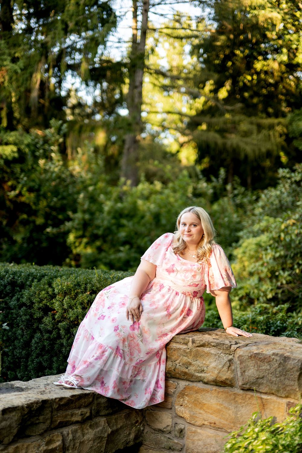 Blonde woman in pink floral maxi dress poses on stone wall in a scenic garden.