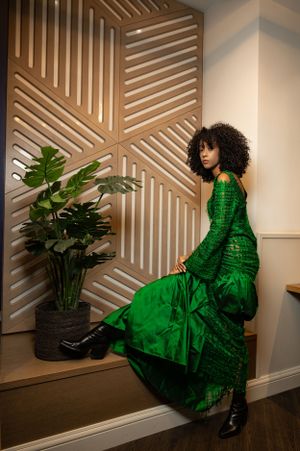 Fashion model in emerald green textured dress poses against geometric wall pattern with indoor plant.
