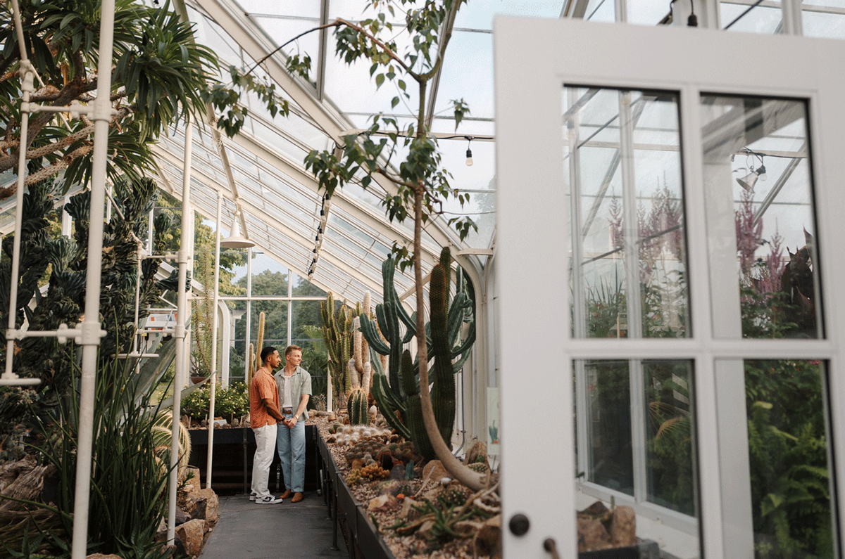 Couple standing together in a lush greenhouse surrounded by cacti and tropical plants with glass ceiling.