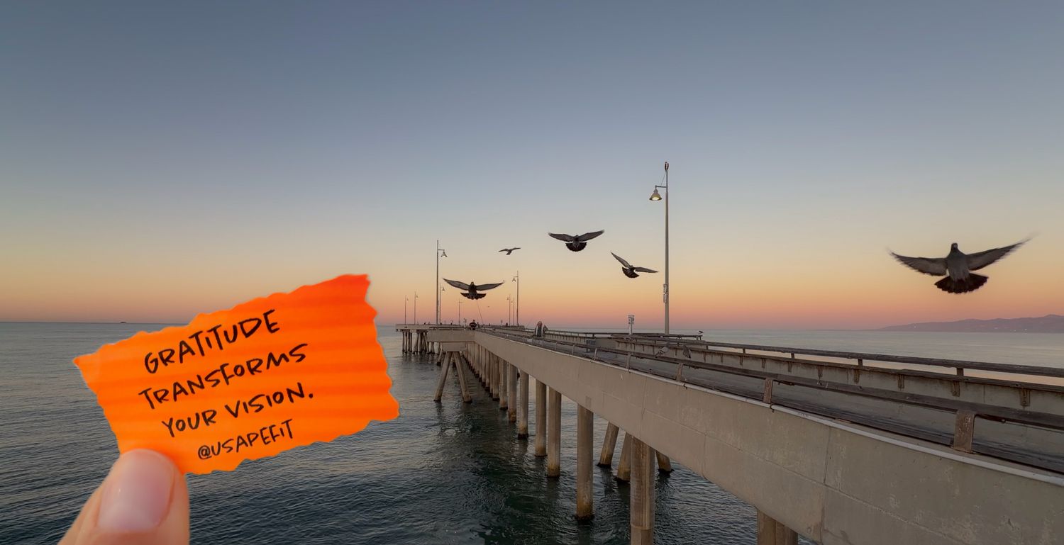 Hand holding an orange gratitude card at sunset pier with birds flying over calm water in golden hour light.