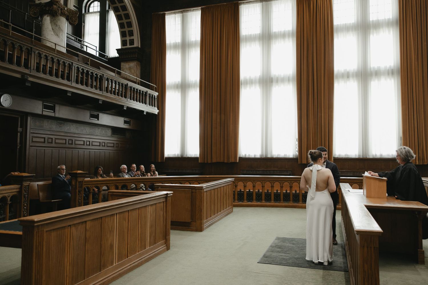 A grand courtroom interior with wooden furnishings, high windows, and a woman in white standing before a judge.