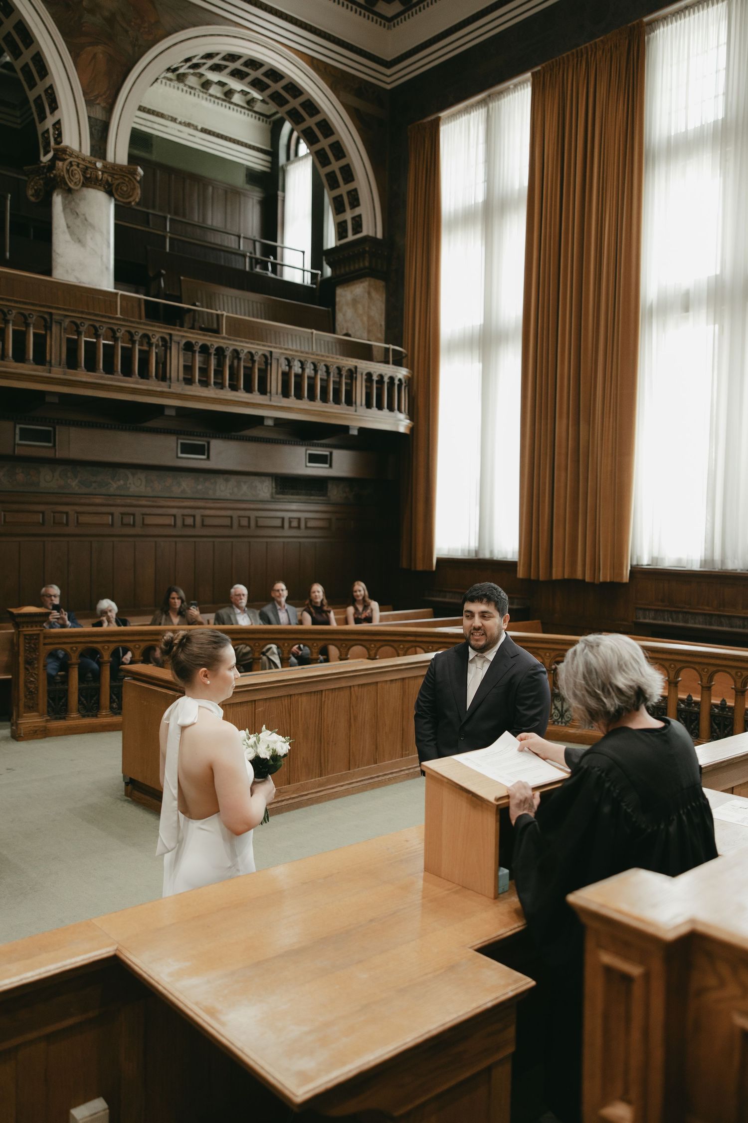 Couple exchanges vows in elegant wood-paneled courthouse wedding ceremony with officiant and guests seated in gallery.