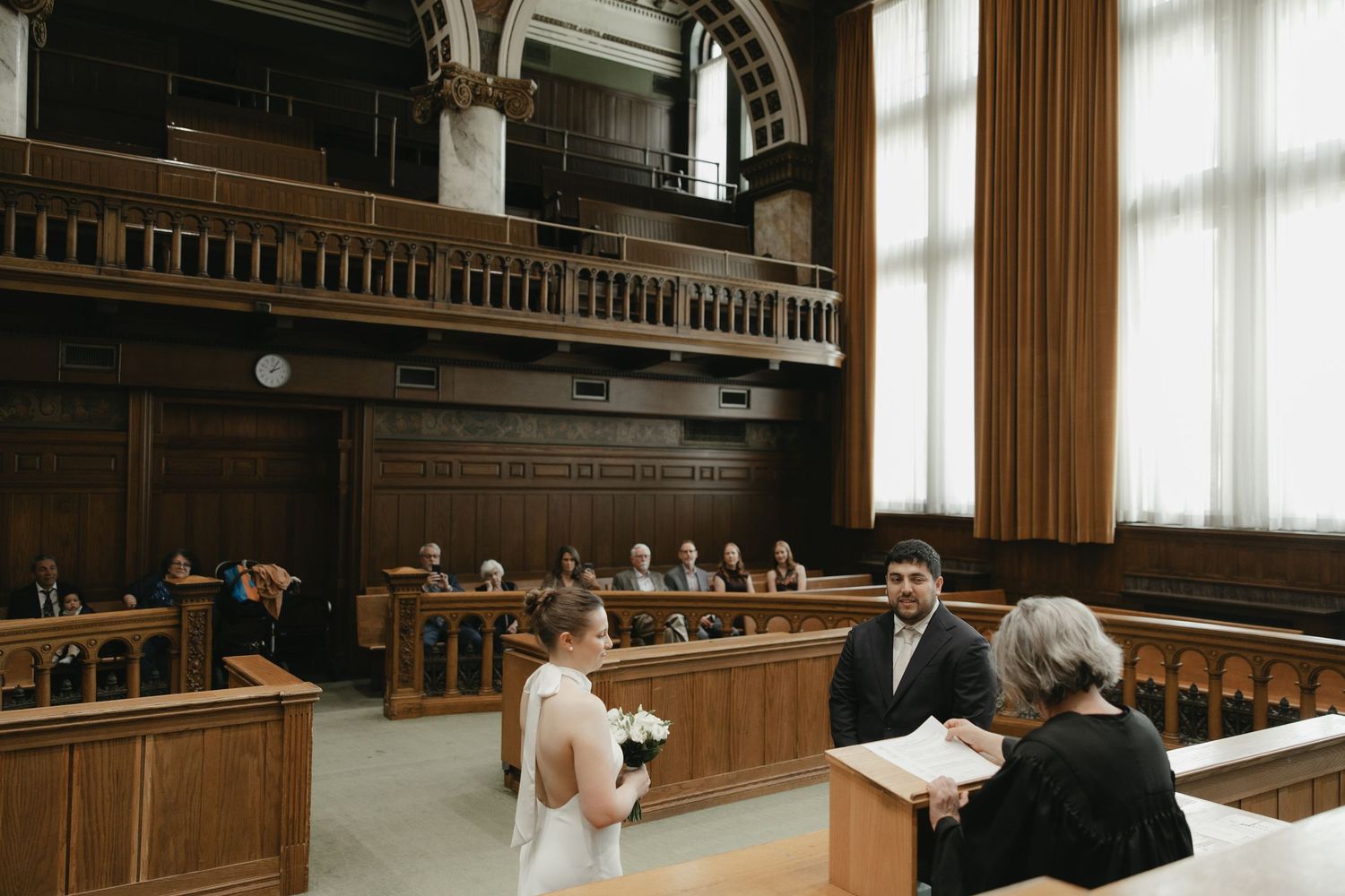 Couple exchanging wedding vows in an elegant courtroom ceremony with wooden architecture and guests seated in the background.