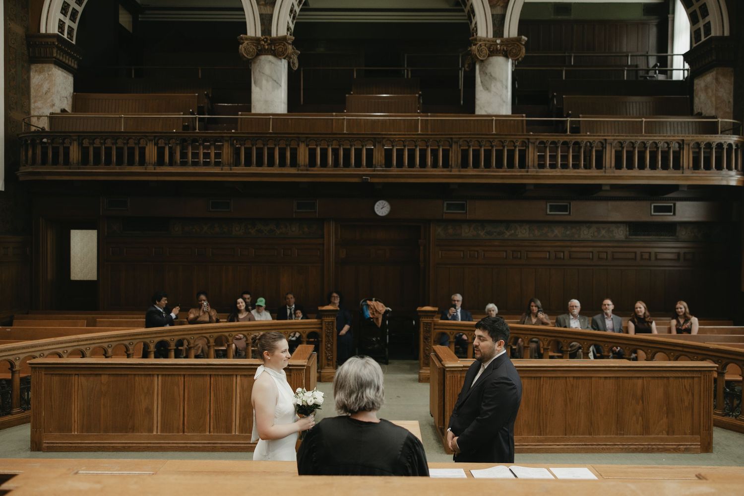 Couple exchanging vows in a grand wooden courtroom with guests seated in jury box during intimate wedding ceremony.