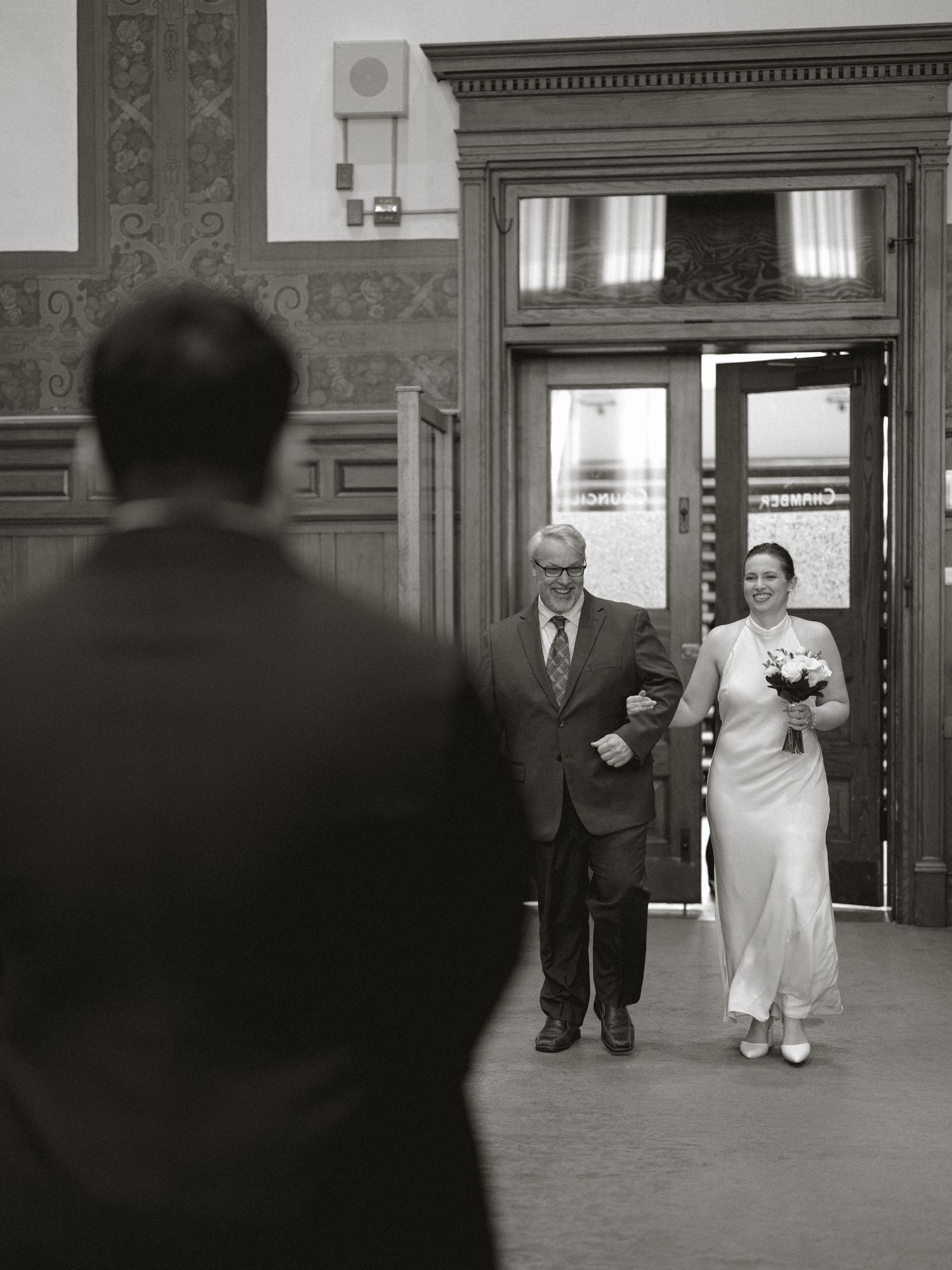 Black and white photo of bride walking down aisle with father toward waiting groom in historic venue.
