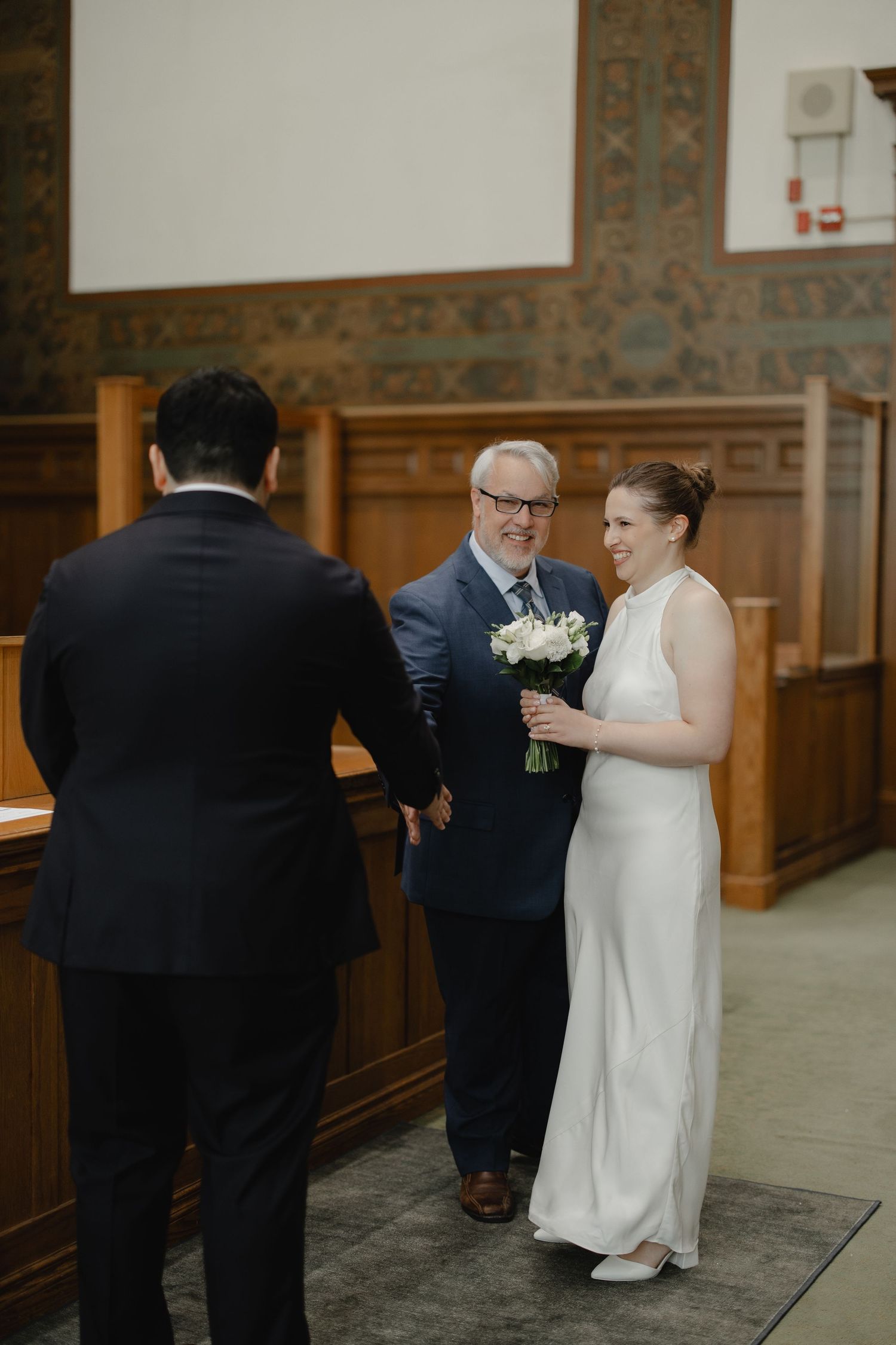 Bride in white dress holds bouquet while smiling at groom during intimate civil wedding ceremony in elegant wood-paneled room.