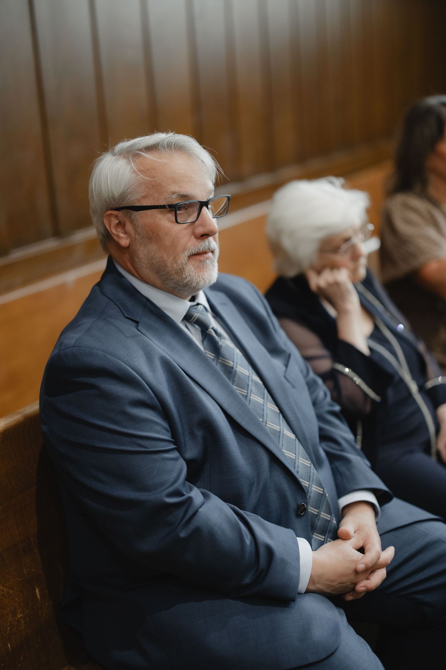 Elderly man in grey suit with glasses sits attentively in wooden pew during wedding ceremony.