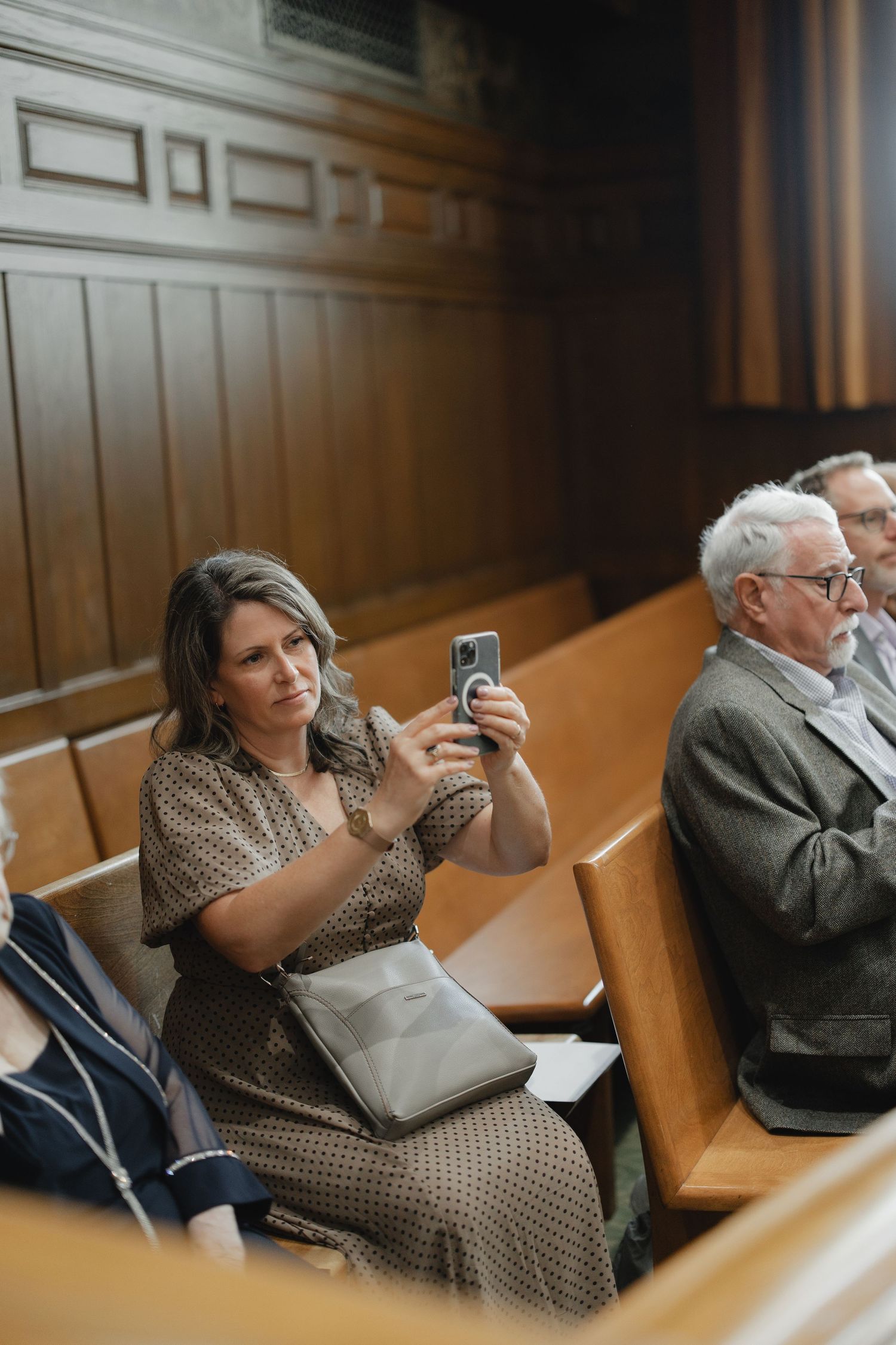 Woman in brown dress photographs a wedding ceremony on her smartphone while seated in wooden church pews.