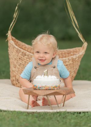 Smiling baby boy in overalls celebrates first birthday with white cake outdoors near wicker basket.