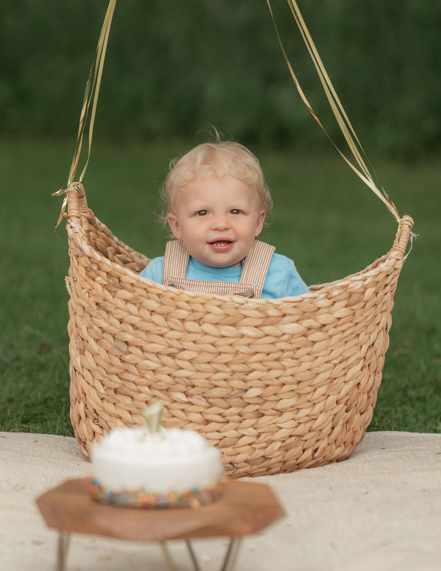 Smiling blonde toddler sitting in a wicker basket with gold straps, with a small white birthday cake in the foreground.