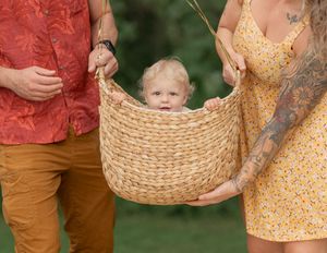 A toddler sits inside a woven basket held by two adults outdoors, one in a floral shirt and one in a yellow dress.