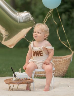 Baby's first birthday cake smash outdoors with ONE romper, silver balloon, and colorful sprinkle cake on wooden stand.