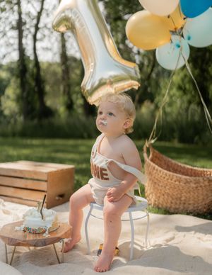 Baby celebrating first birthday outdoors with number one balloon, colorful balloons, and smash cake on sand.