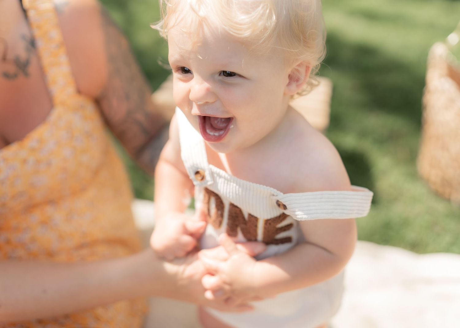Joyful baby with curly blonde hair smiling and holding a cake topper during a first birthday cake smash outdoors.