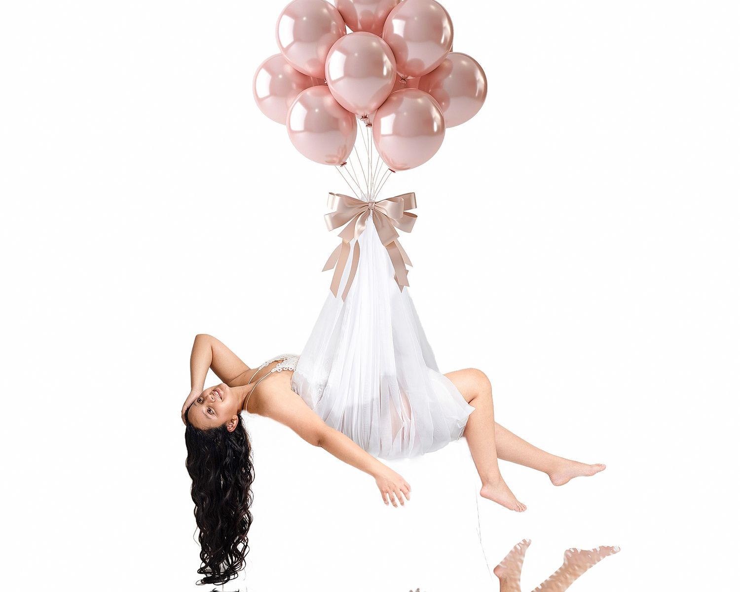 Woman in white dress posed as if floating, held up by pink and rose gold metallic balloons with a bow.