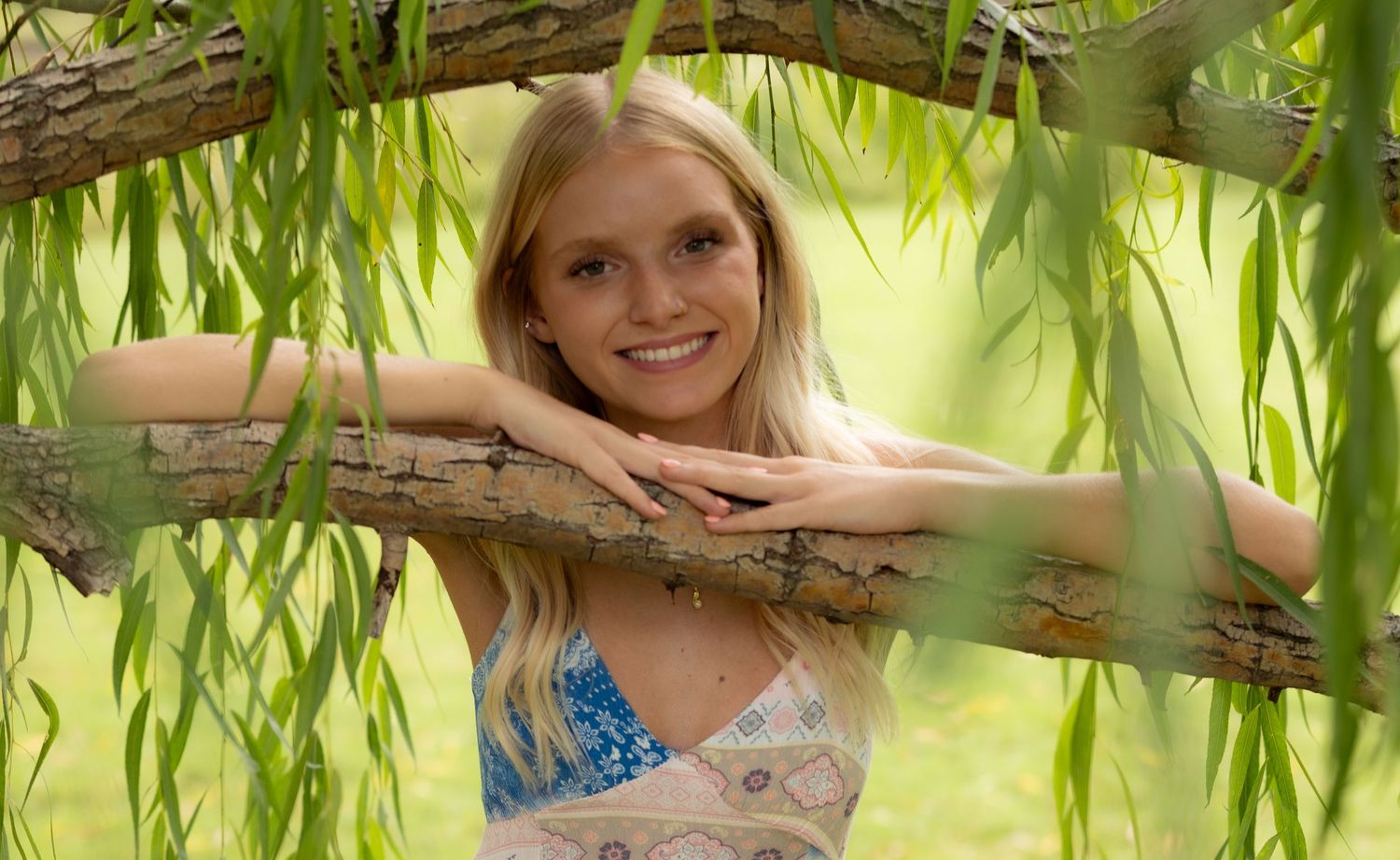 Smiling blonde woman leaning on a tree branch surrounded by lush green willow leaves outdoors.