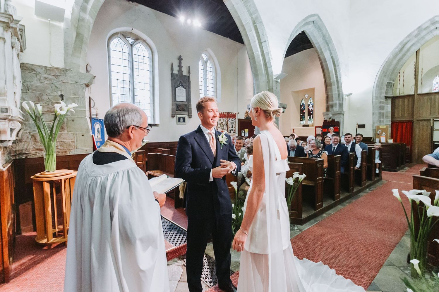 Bride and groom exchange vows with officiant inside a traditional stone church decorated with white calla lilies.