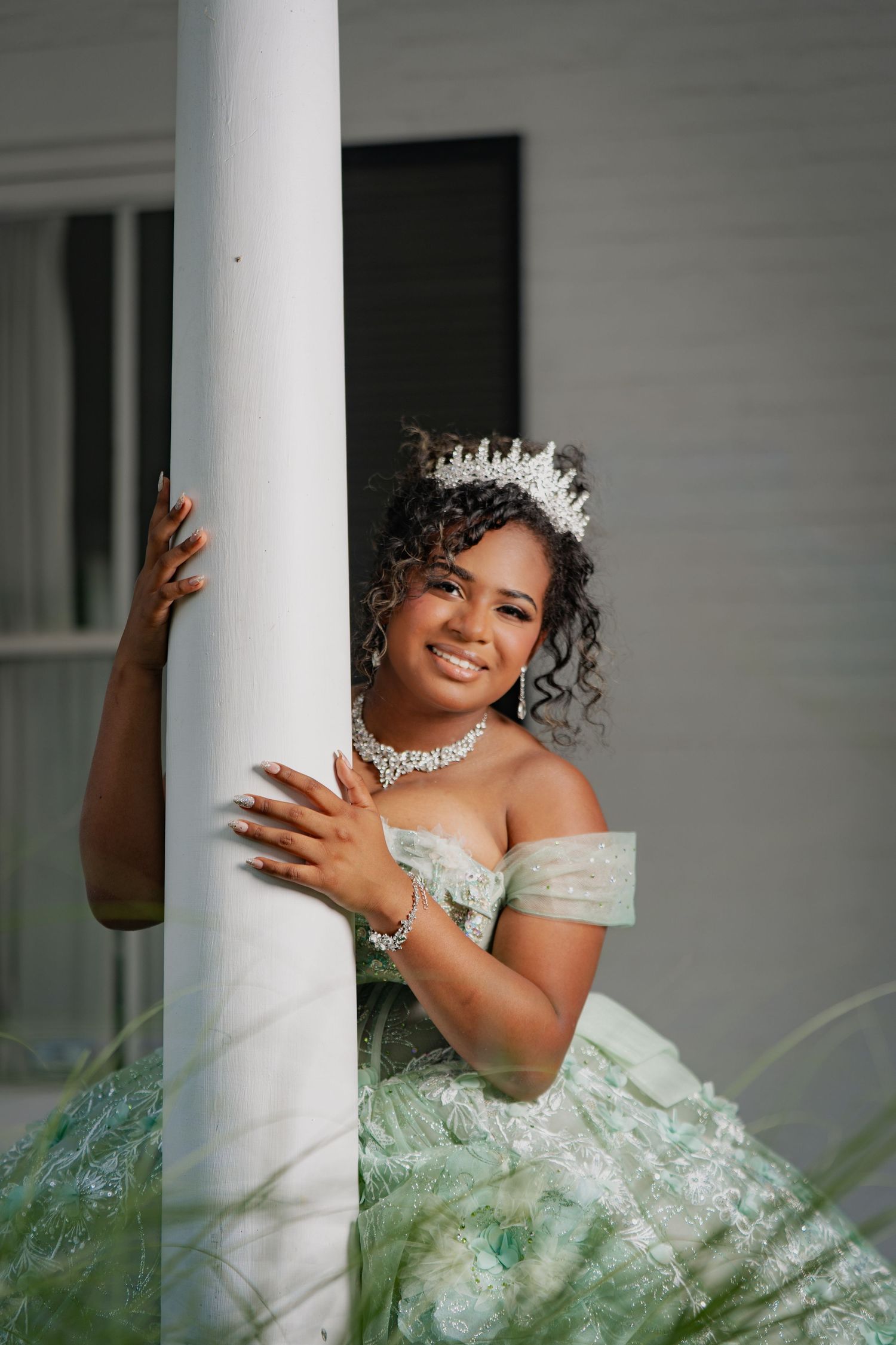 Young woman in pink quinceañera ball gown with tiara joyfully raises floral bouquet outdoors.