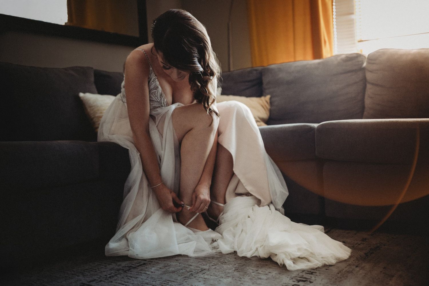 Bride in a beaded Maggie Sottero gown buckling her shoes on a sofa in window light