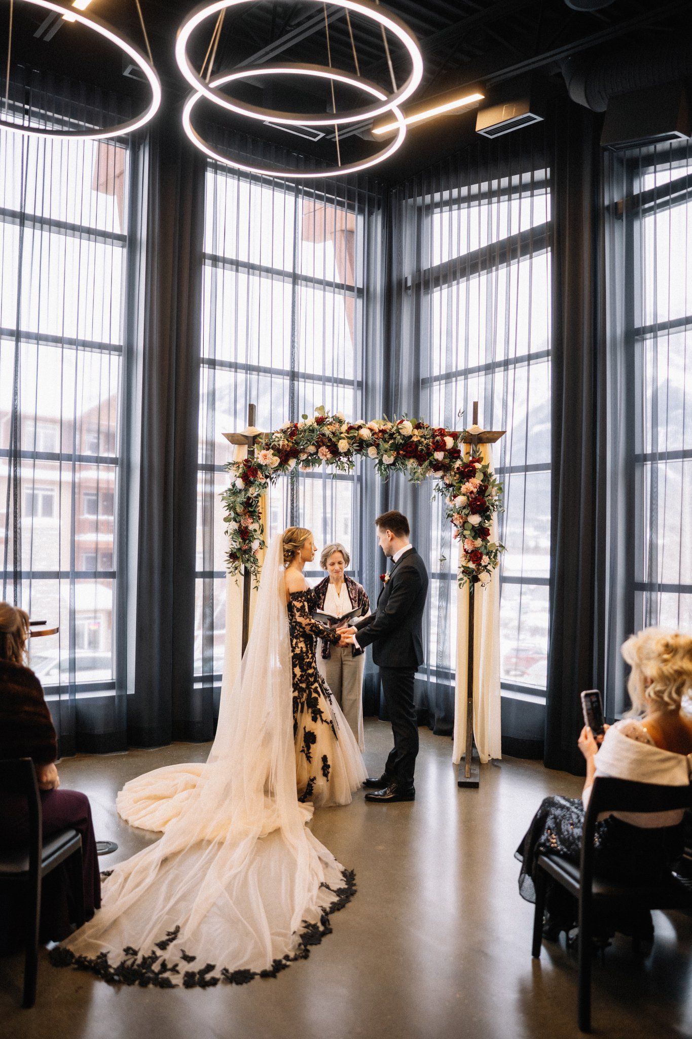 Bride and groom at a red rose floral arch at Mainspace, her long black lace-trimmed train spread across the floor
