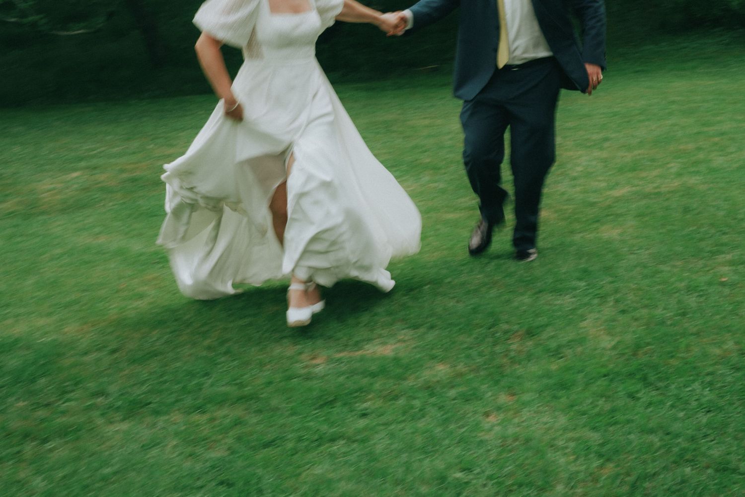 Bride in white gown and groom in navy suit holding hands running across lush green grass at outdoor wedding.