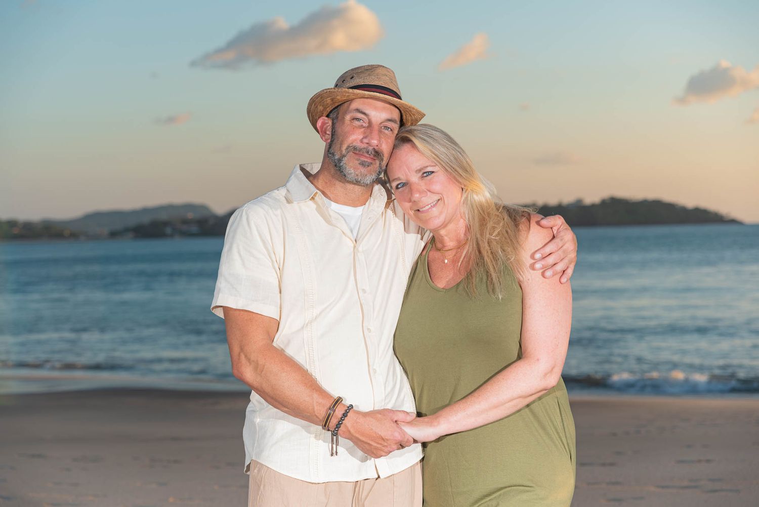Couple embracing on a tropical beach at sunset, man in straw hat and white shirt, woman in green dress.