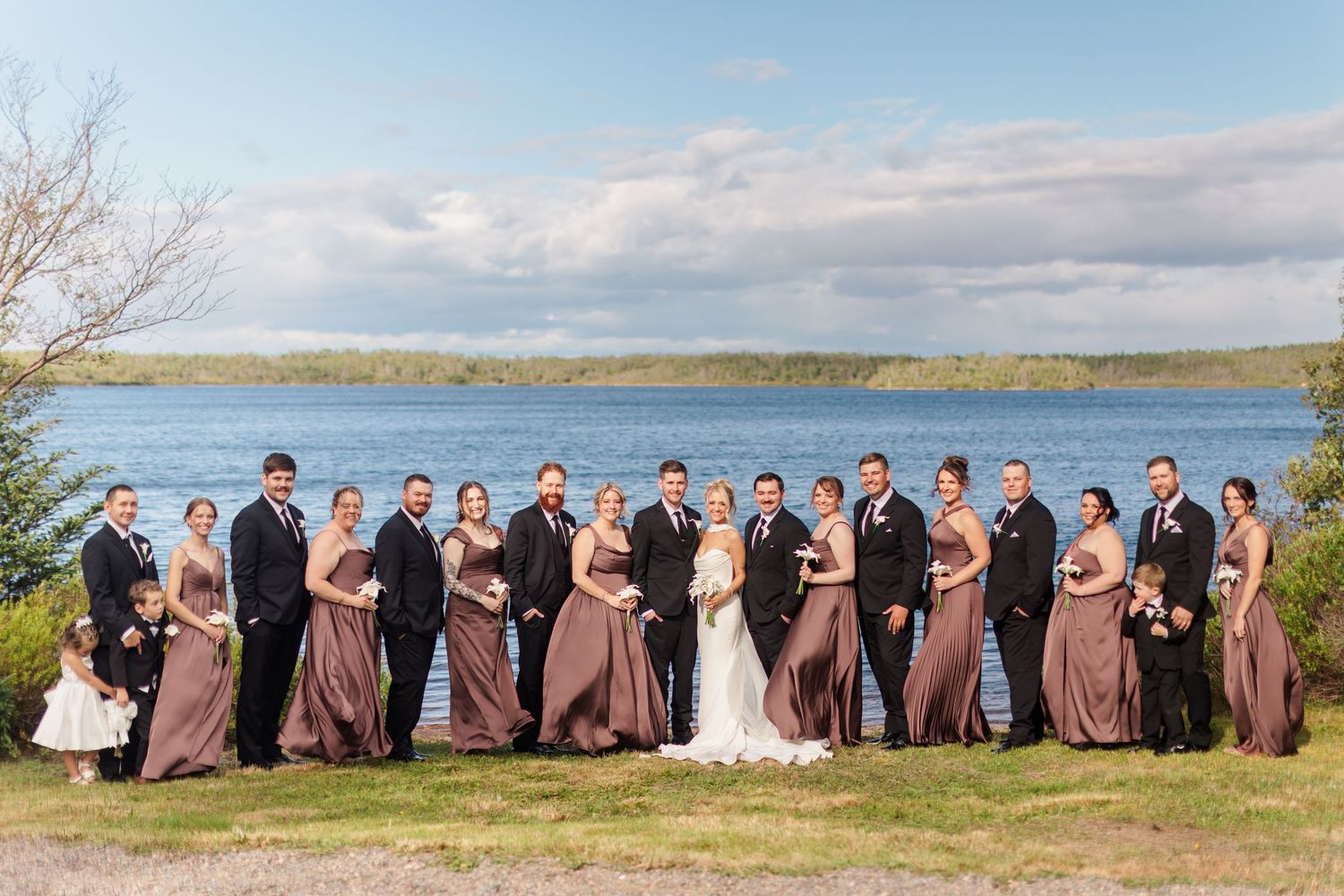 Large wedding party poses by a lakeside on a sunny day, bridesmaids in mauve gowns and groomsmen in black suits.