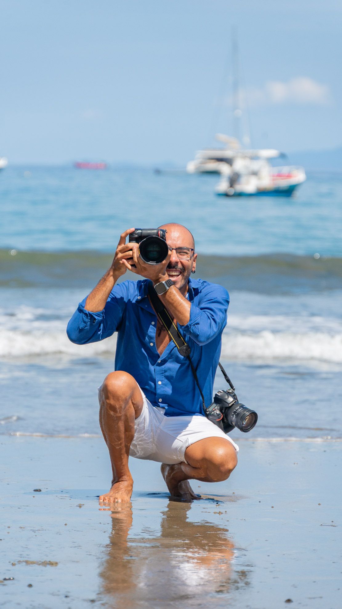 Bald man in blue shirt crouching on beach, holding camera up to photograph, with ocean and boats behind him.