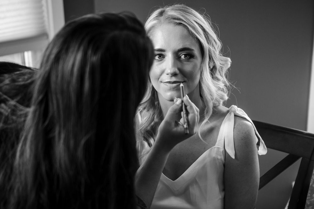 Black and white photo of a makeup artist applying lipstick to a smiling blonde bride during wedding preparations.