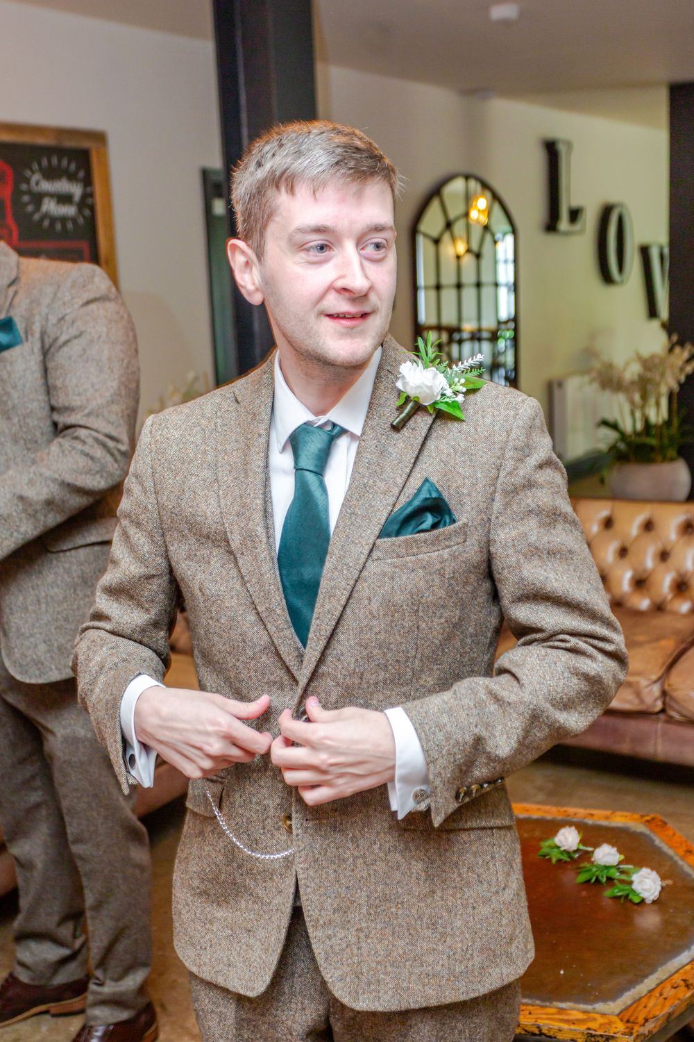 Groom in brown tweed suit with green tie and white boutonniere buttoning jacket at wedding venue.