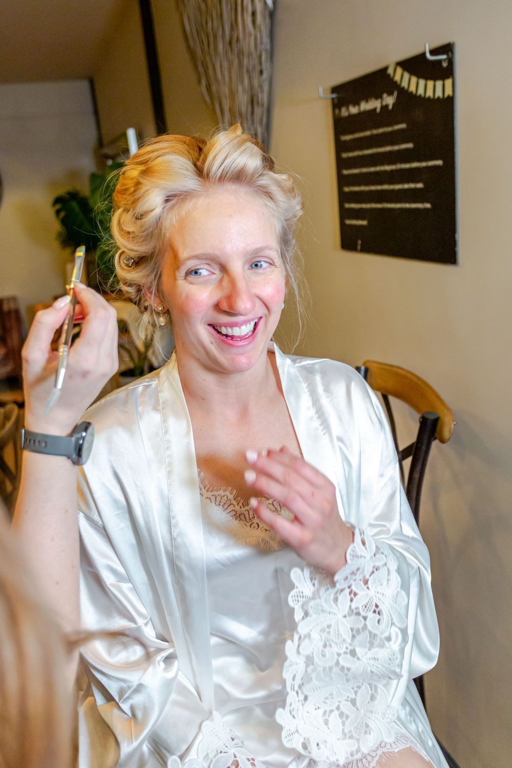 Smiling bride in white satin robe gets makeup applied while preparing for her wedding day.