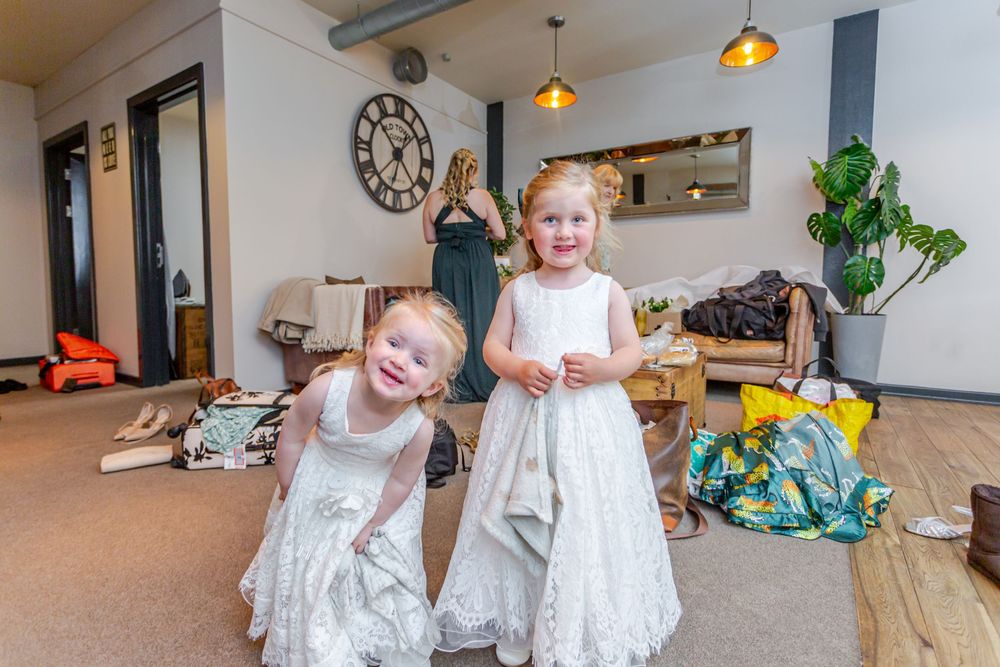 Two young flower girls in white lace dresses smile in a decorated room during wedding preparations.