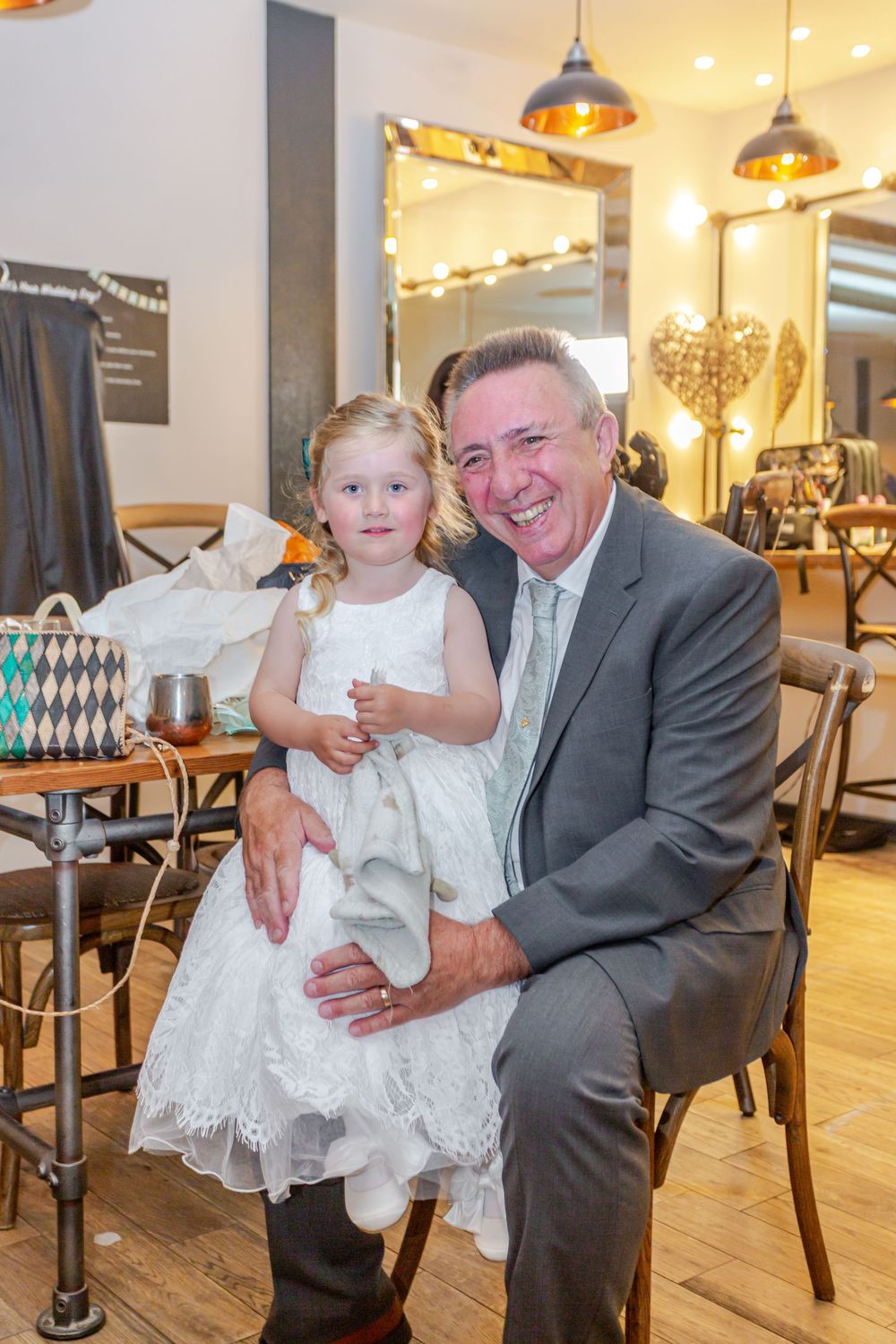 Elderly man in gray suit holds young girl in white dress at a wedding reception venue.
