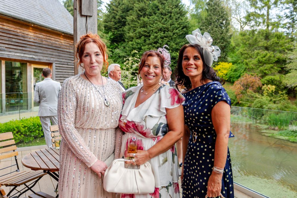 Three women in elegant dresses and fascinators posing together at an outdoor garden wedding.