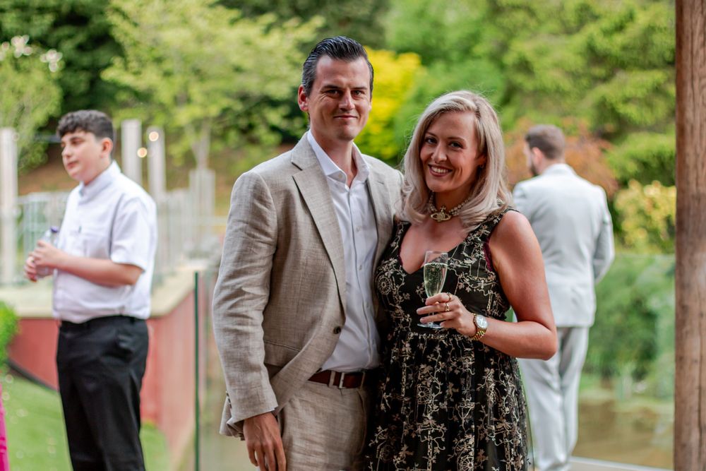 A smiling couple poses outdoors at a garden party, woman holding champagne, guests mingling in background.