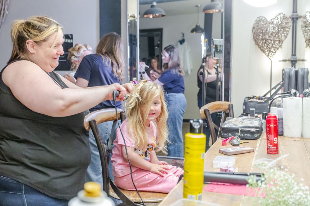 Woman styling young girl's hair at a salon mirror with beauty products on the counter.
