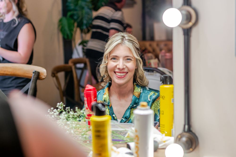 Smiling blonde woman sitting at a vanity mirror with bulb lights, surrounded by hair products in a bright room.