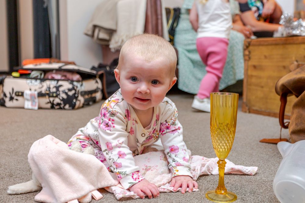 Smiling baby in floral pajamas on carpet next to a yellow glass goblet, with another child in background.