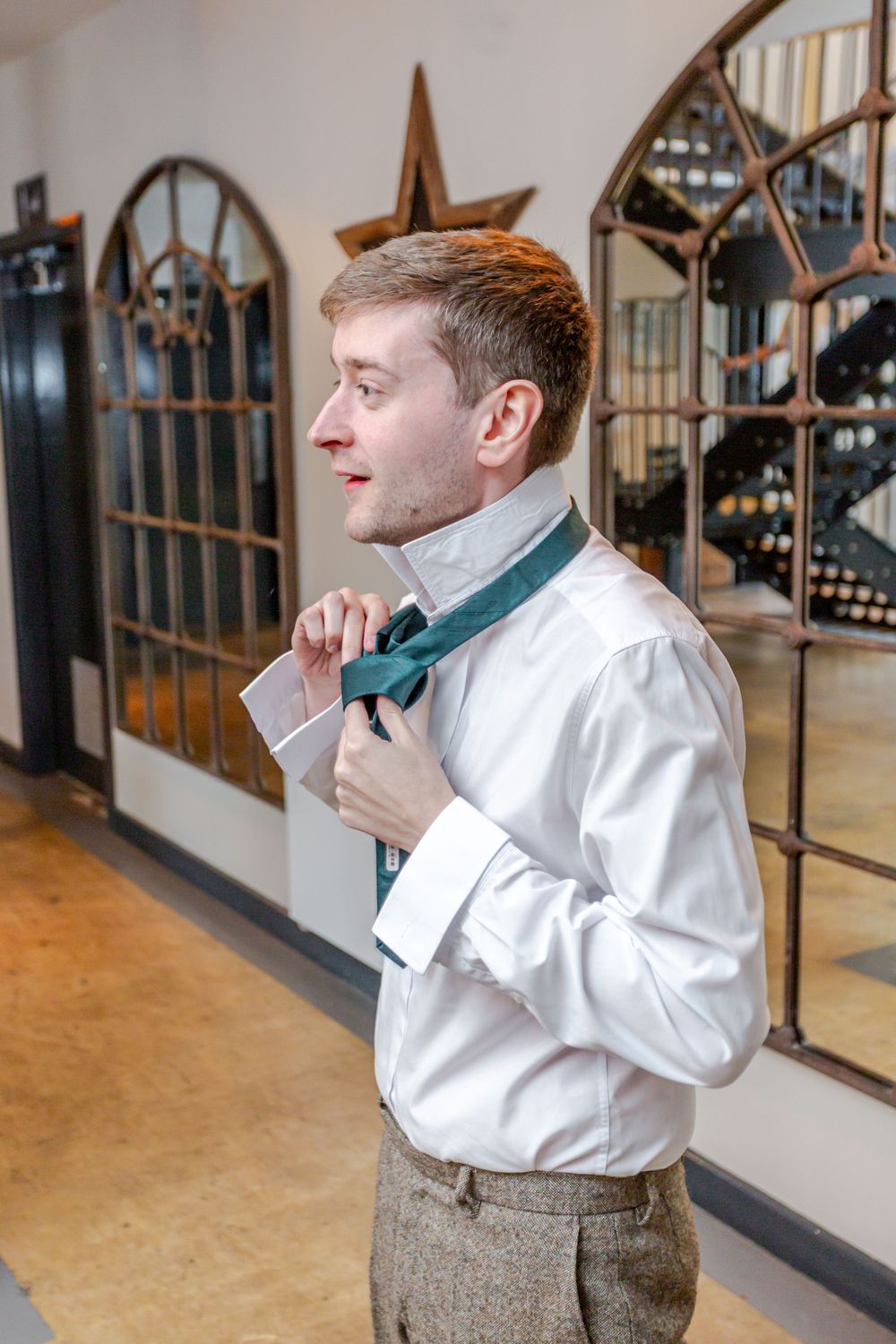 Groom adjusting green bow tie in white shirt and tweed trousers before wedding ceremony.