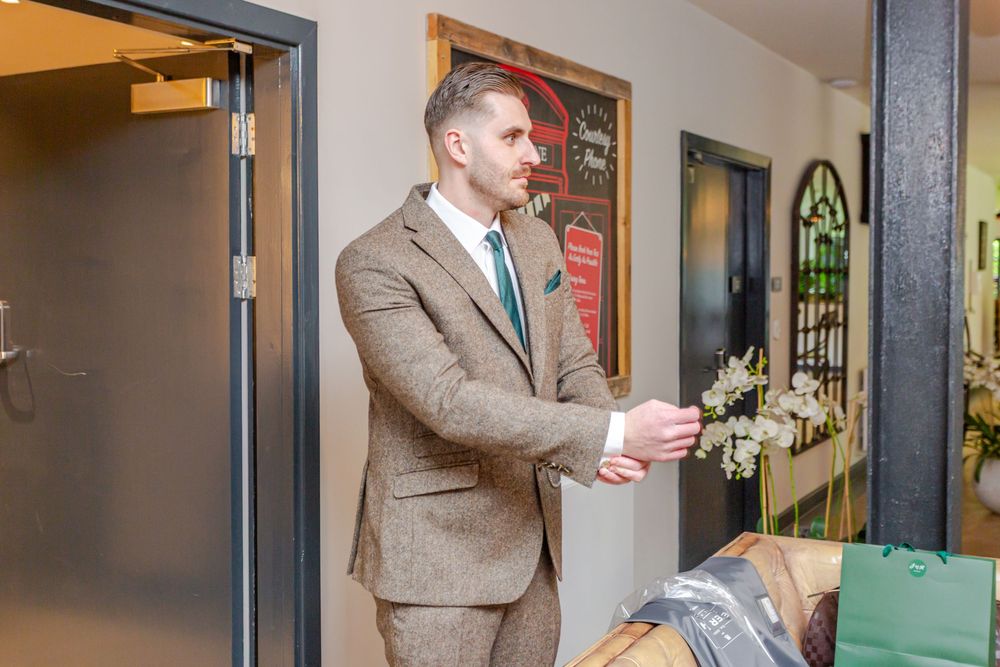 Man in brown tweed suit adjusting cufflinks indoors near elevator, holding white flowers at a formal event.