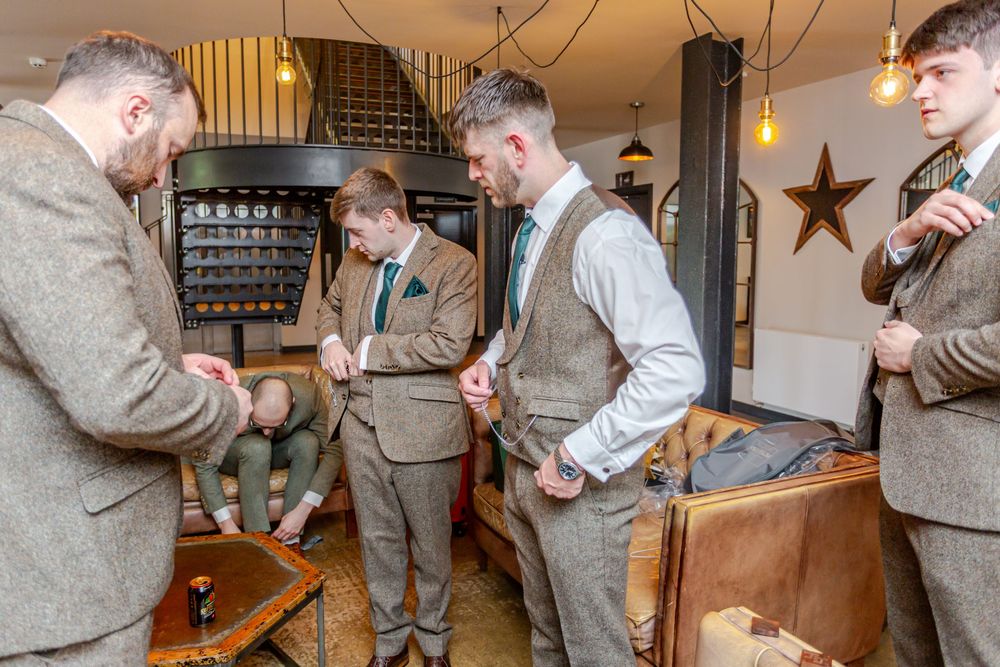 Groomsmen in tweed suits preparing for a wedding ceremony in a rustic venue with vintage decor.