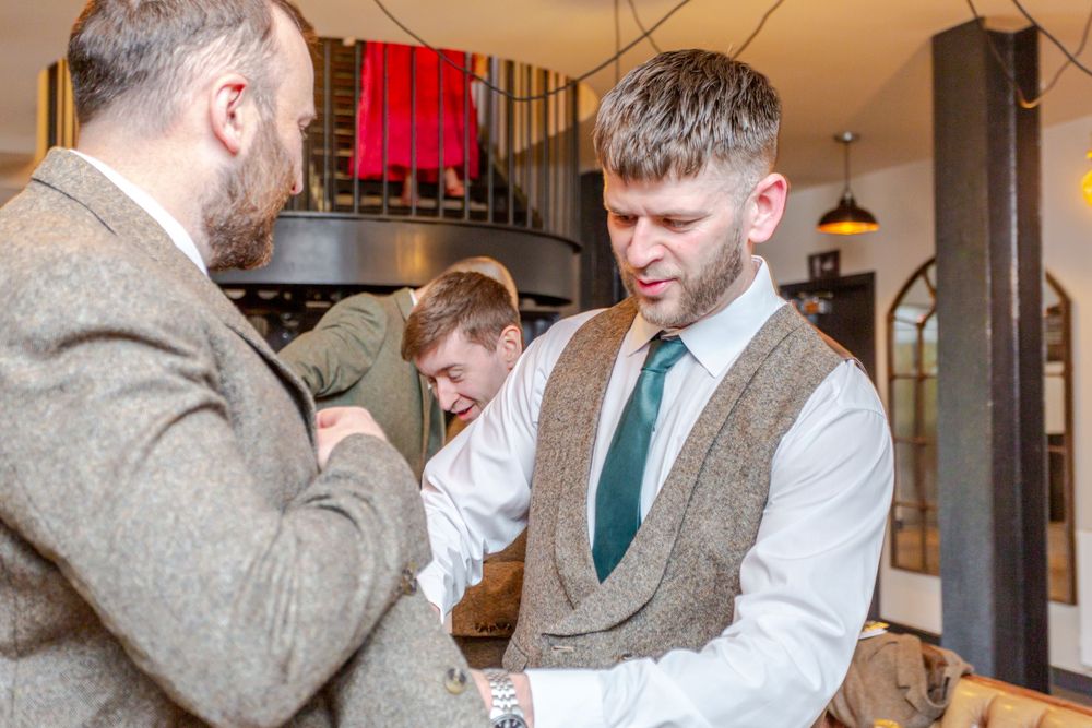 Two men in tweed suits and ties converse indoors at a wedding venue.
