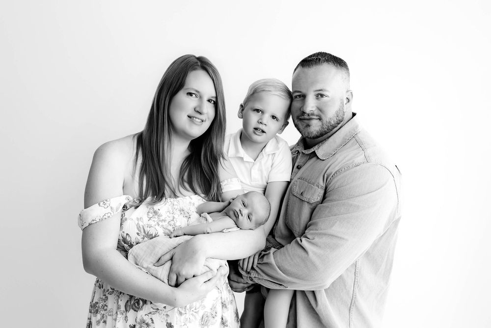 Black and white family portrait with parents holding newborn baby and toddler son in a studio setting.