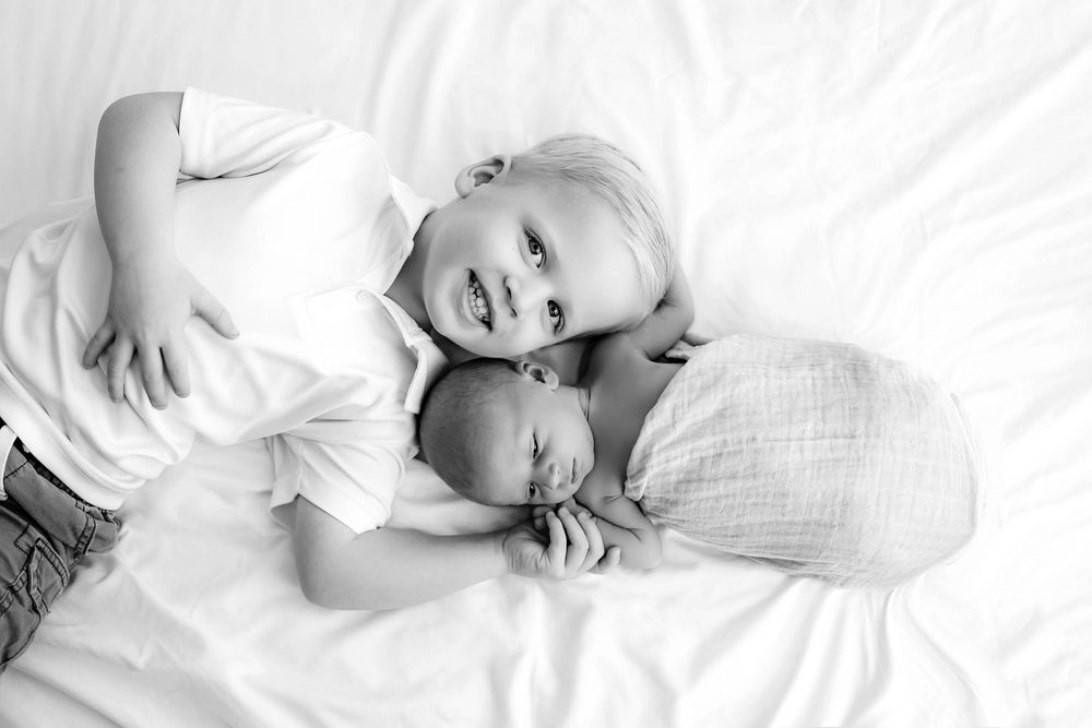 Black and white photo of a young boy lying on a bed smiling while gently holding a sleeping newborn baby.