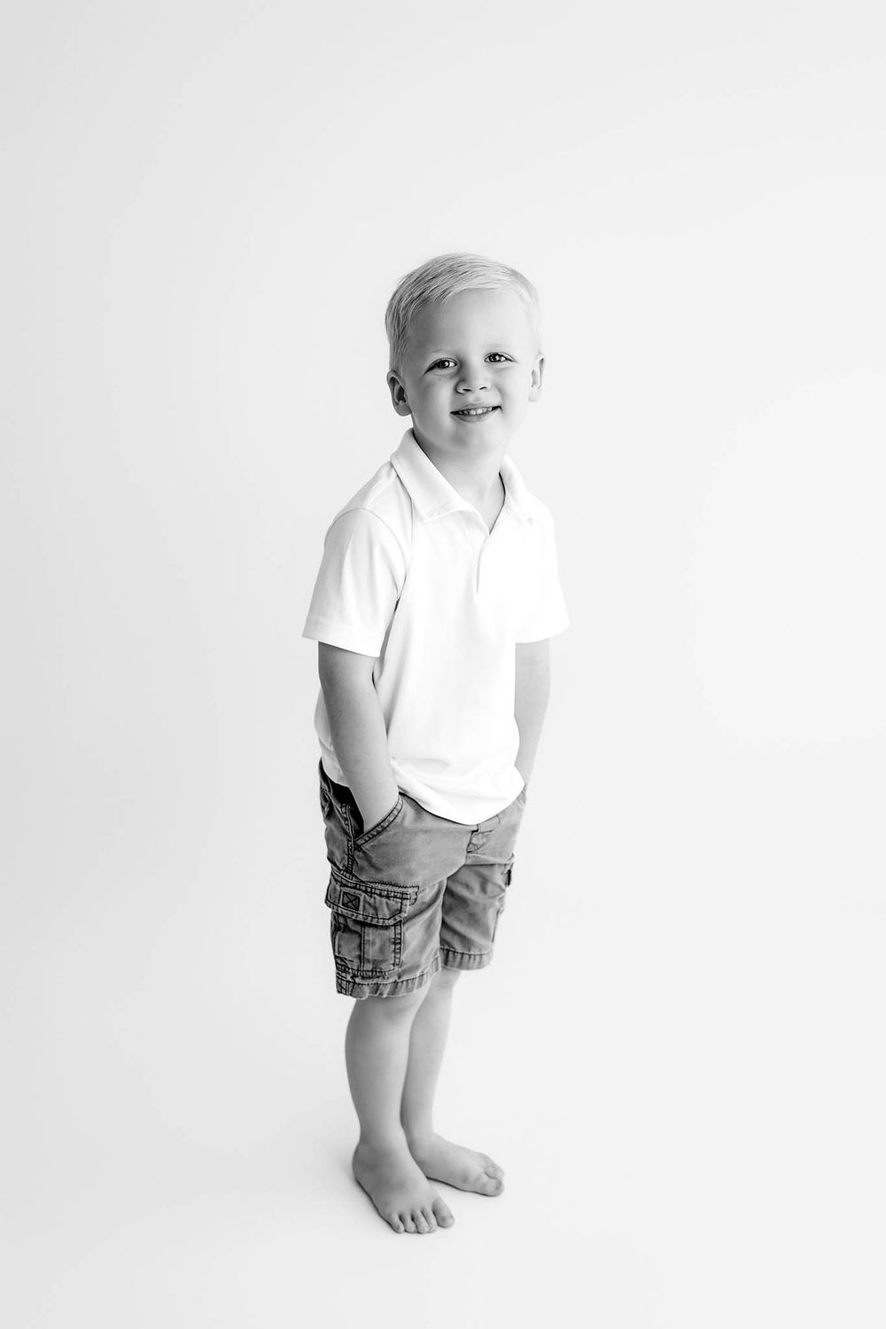 Smiling young boy in white polo shirt and cargo shorts standing barefoot against white background.