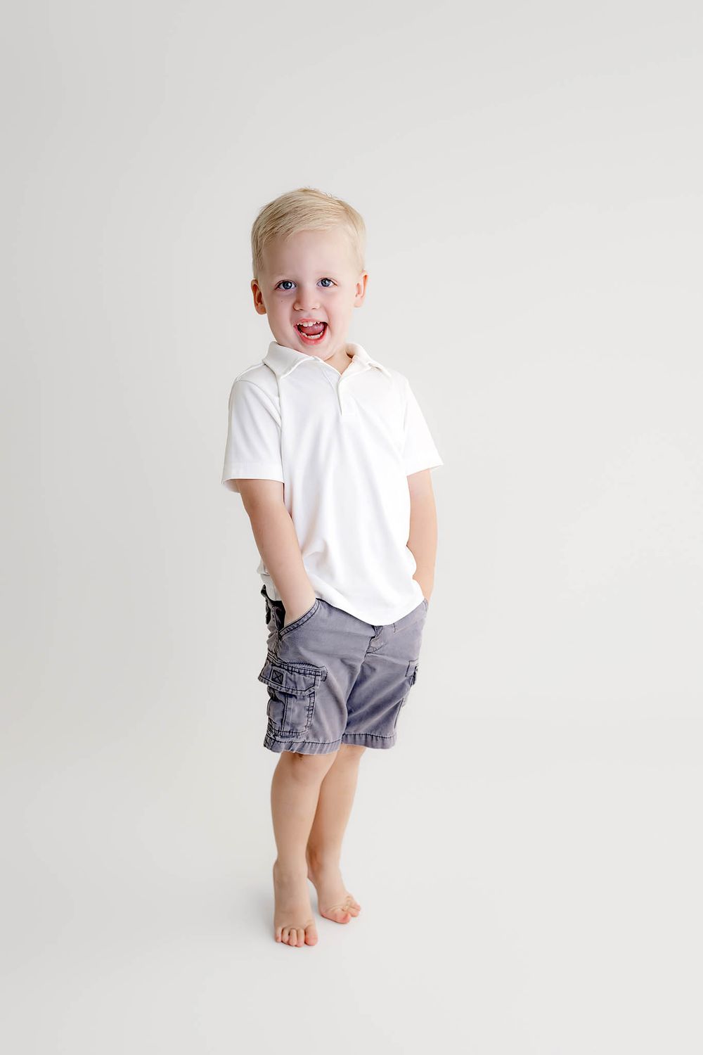 A cheerful blonde toddler boy in a white polo shirt and gray cargo shorts stands barefoot against a white background.