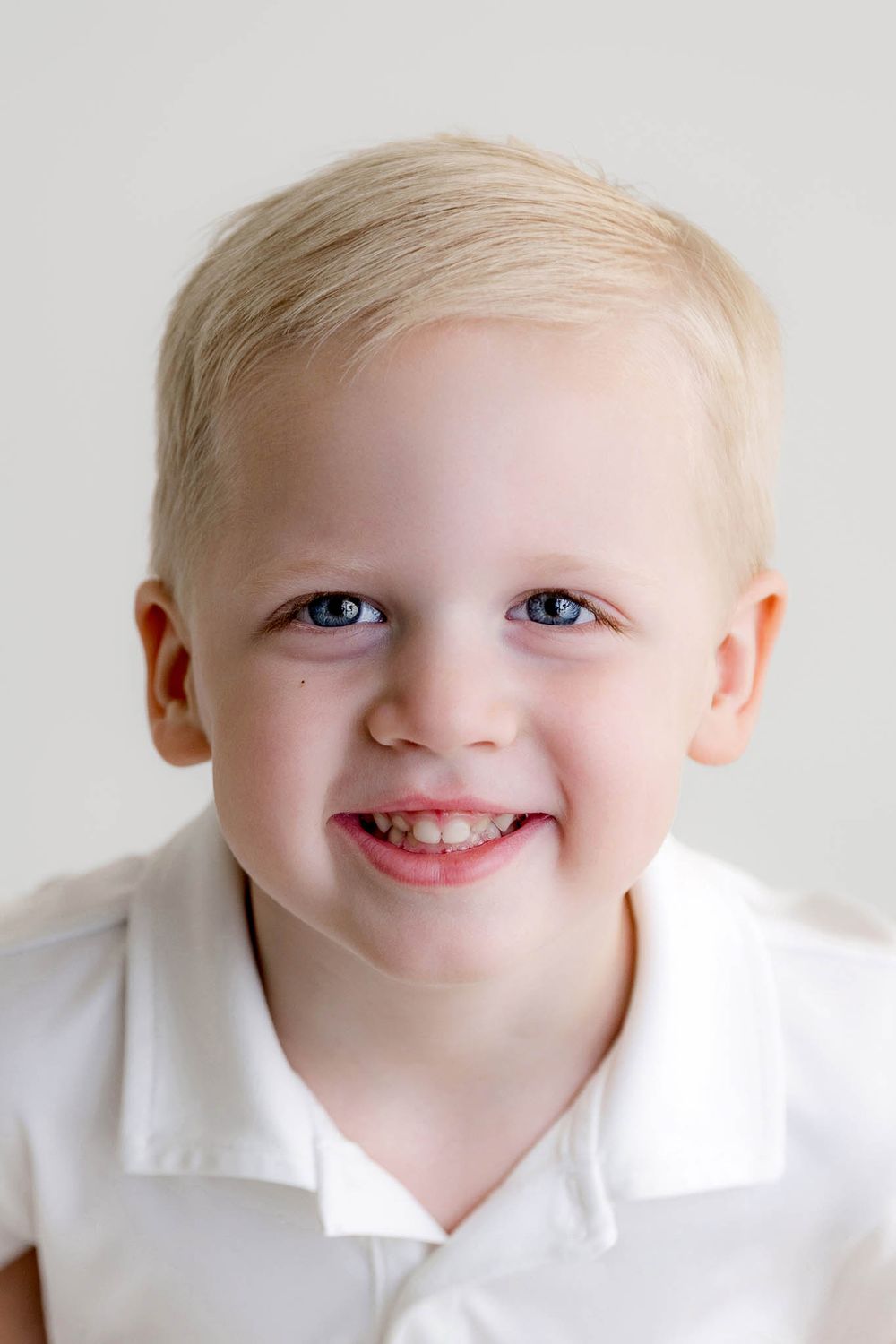 Smiling blonde toddler boy with blue eyes wearing a white polo shirt against a light background.