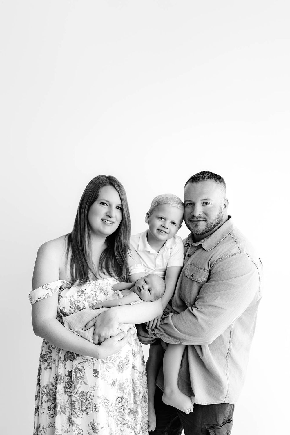 Black and white family portrait with mother holding newborn, father carrying toddler son, studio background.