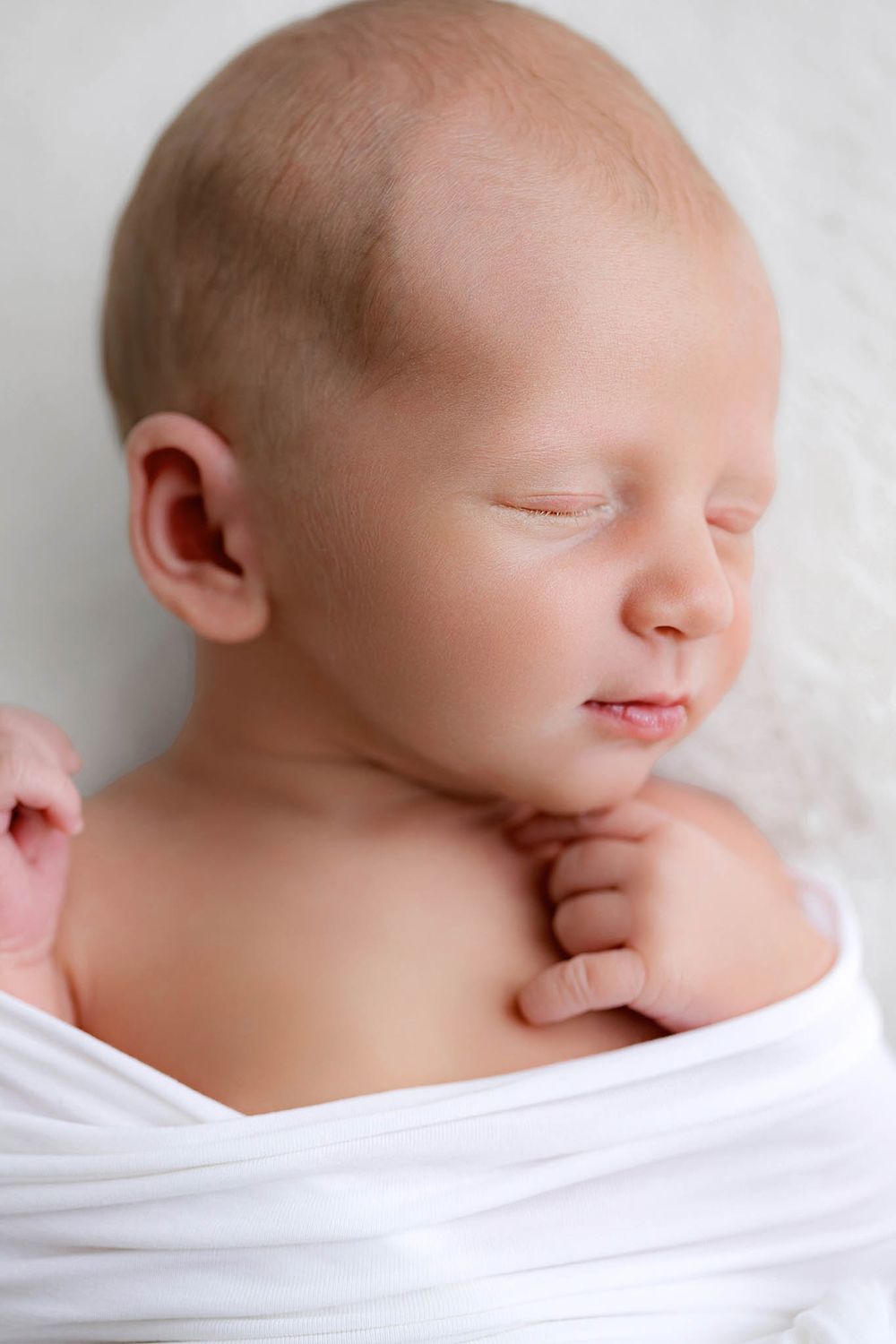 Peaceful newborn baby sleeping wrapped in white cloth with tiny hand visible near chin.