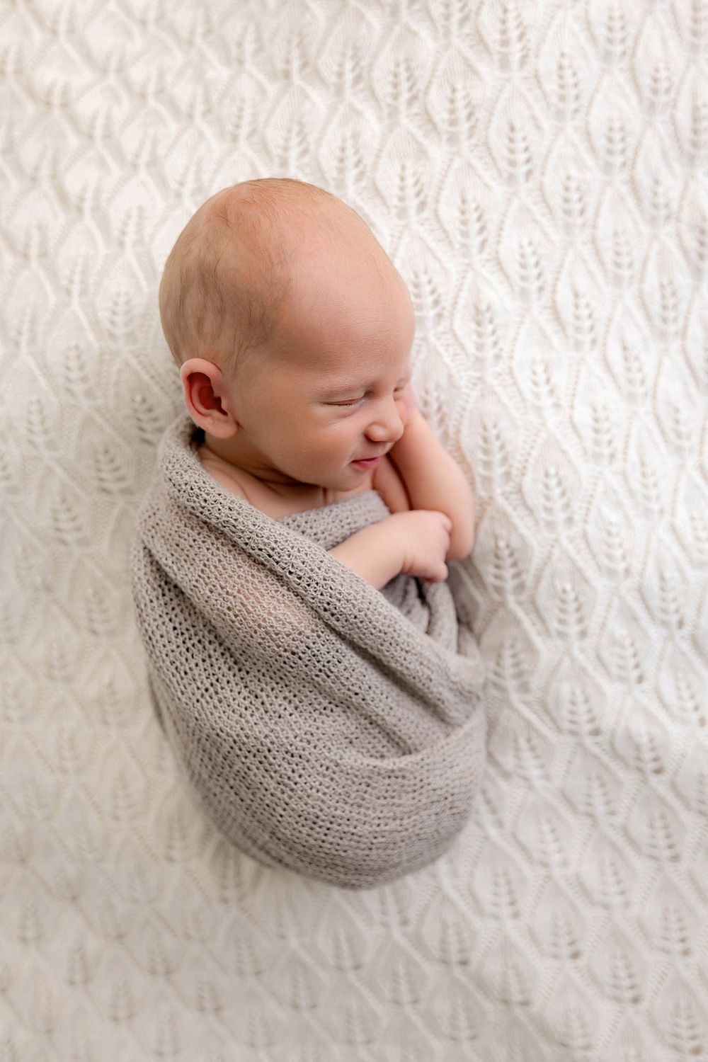 Sleeping newborn baby wrapped in grey knit blanket on white textured surface.
